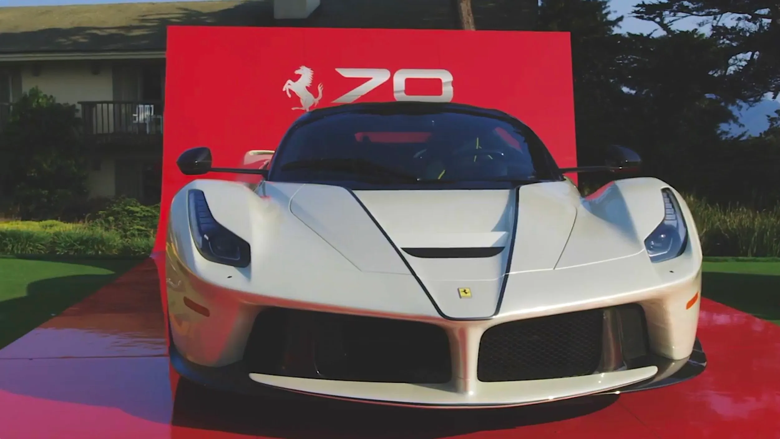 Front view of a white Ferrari sports car displayed on a red platform with a red backdrop showing the Ferrari logo and the number 70.