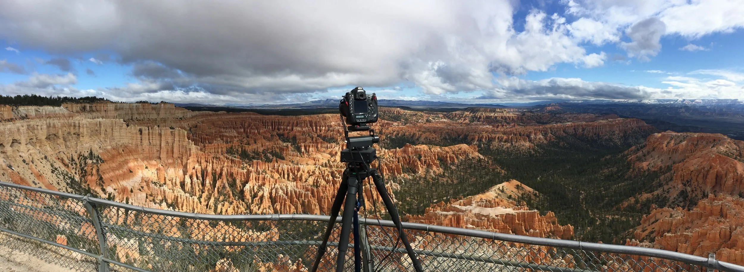 DSLR camera mounted on a tripod facing a vast canyon landscape with orange and beige rock formations under a cloudy sky.