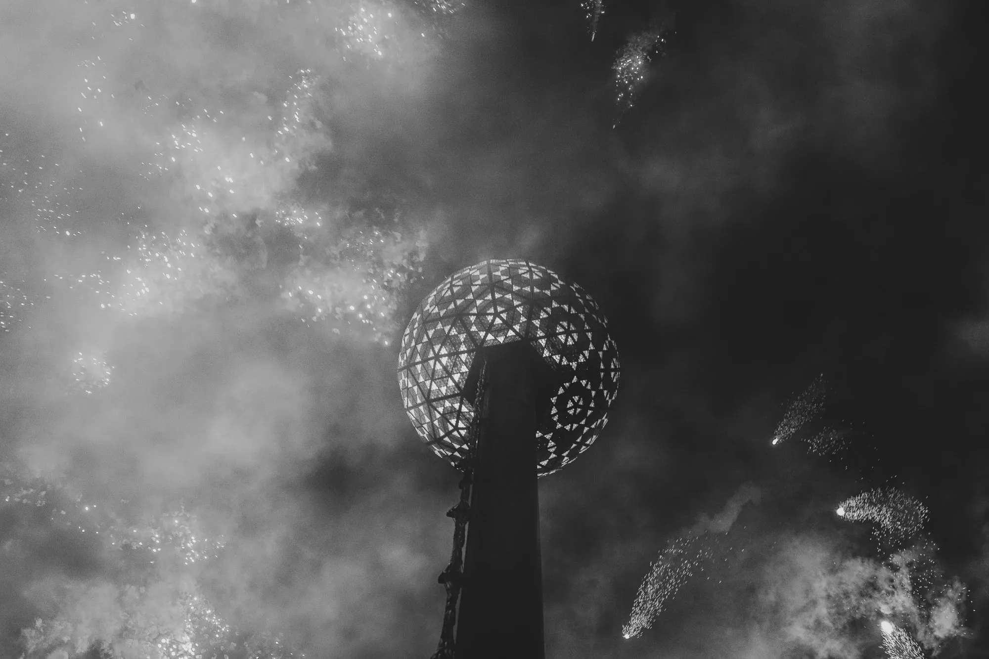 Black and white photo of a tower topped by a glowing sphere amid smoke and fireworks.