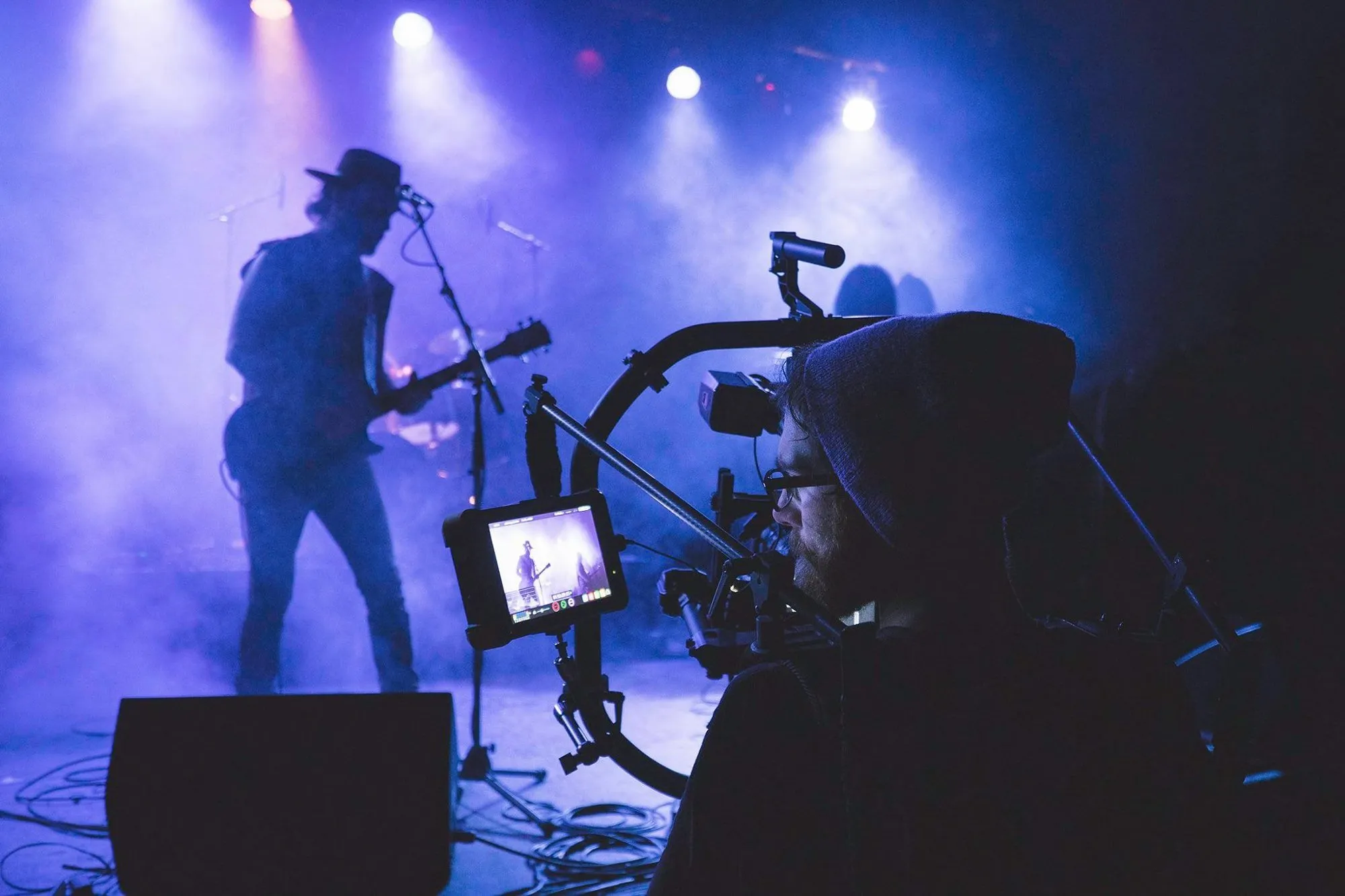 Camera operator filming a musician playing electric guitar and singing on a foggy, blue-lit stage.