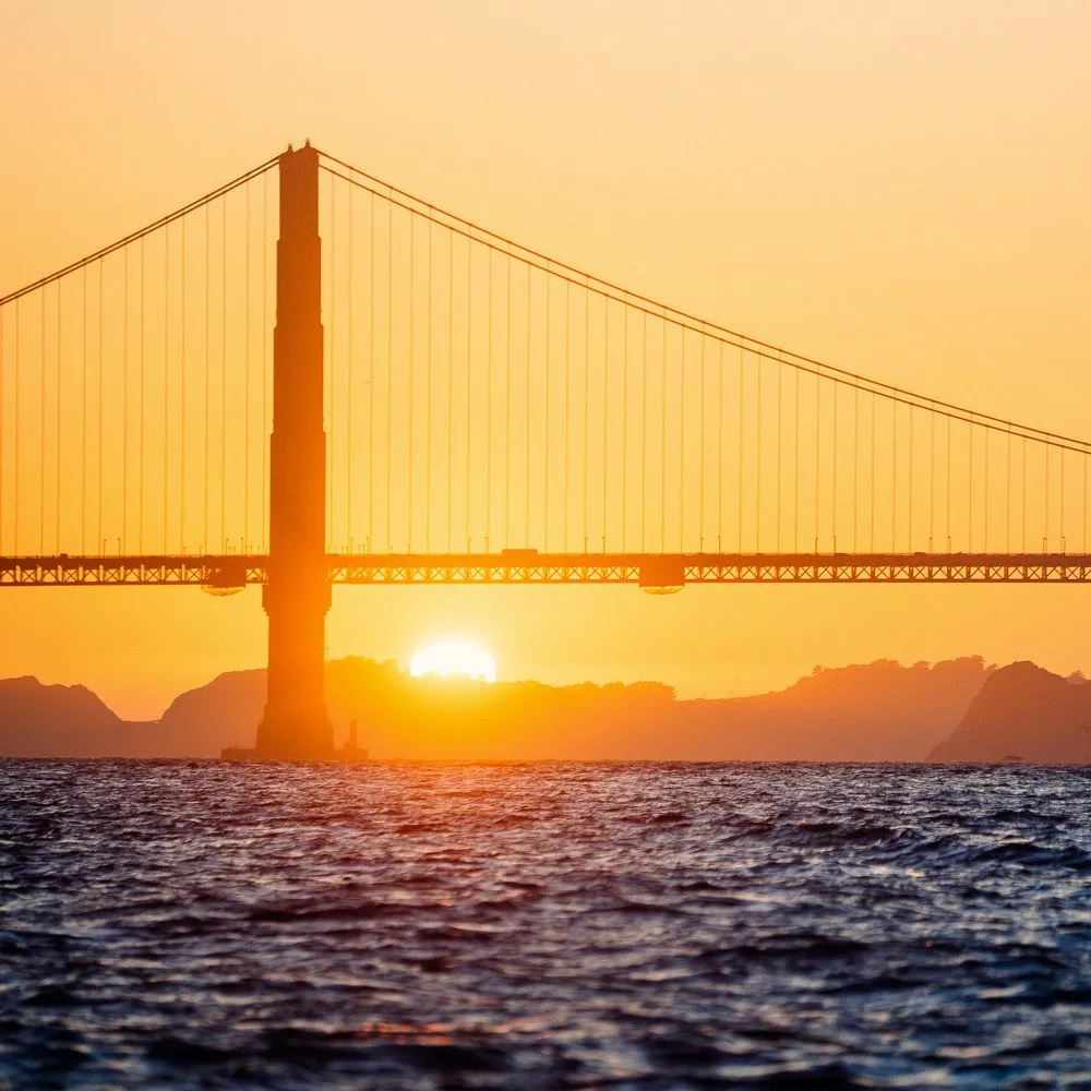 Sun setting behind the Golden Gate Bridge with the bridge silhouetted against an orange sky and water in the foreground.