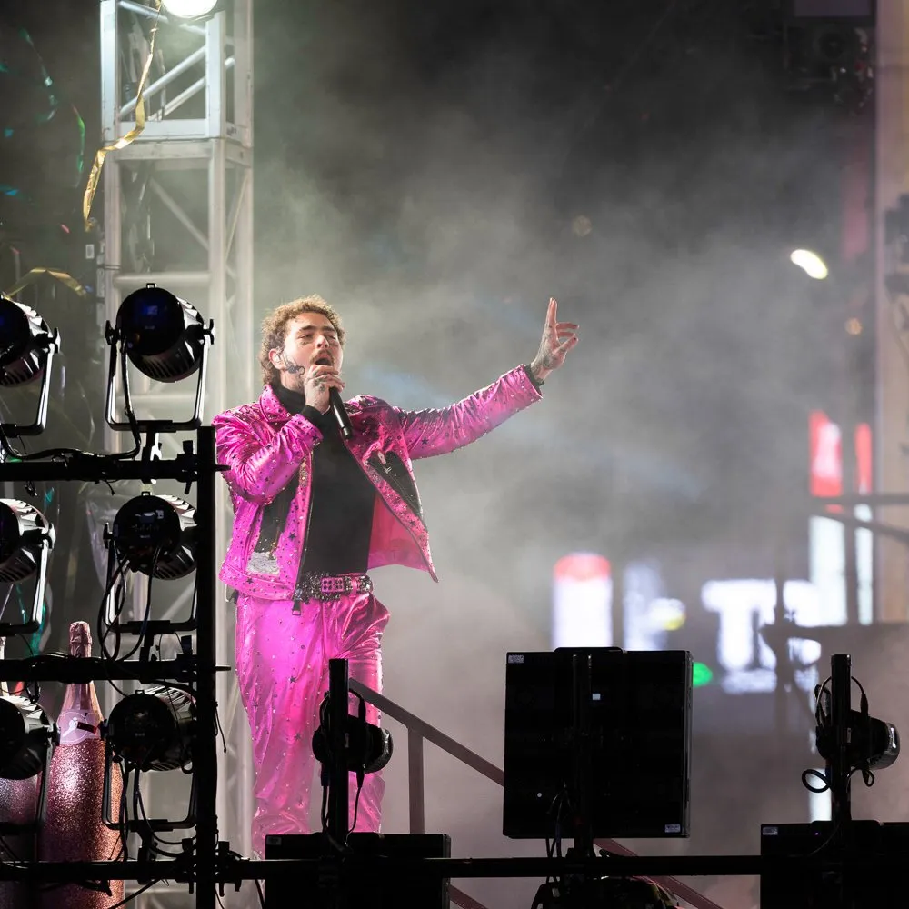 Male performer singing on stage wearing a bright pink glittery suit and holding a microphone, with stage lights and smoke in the background.