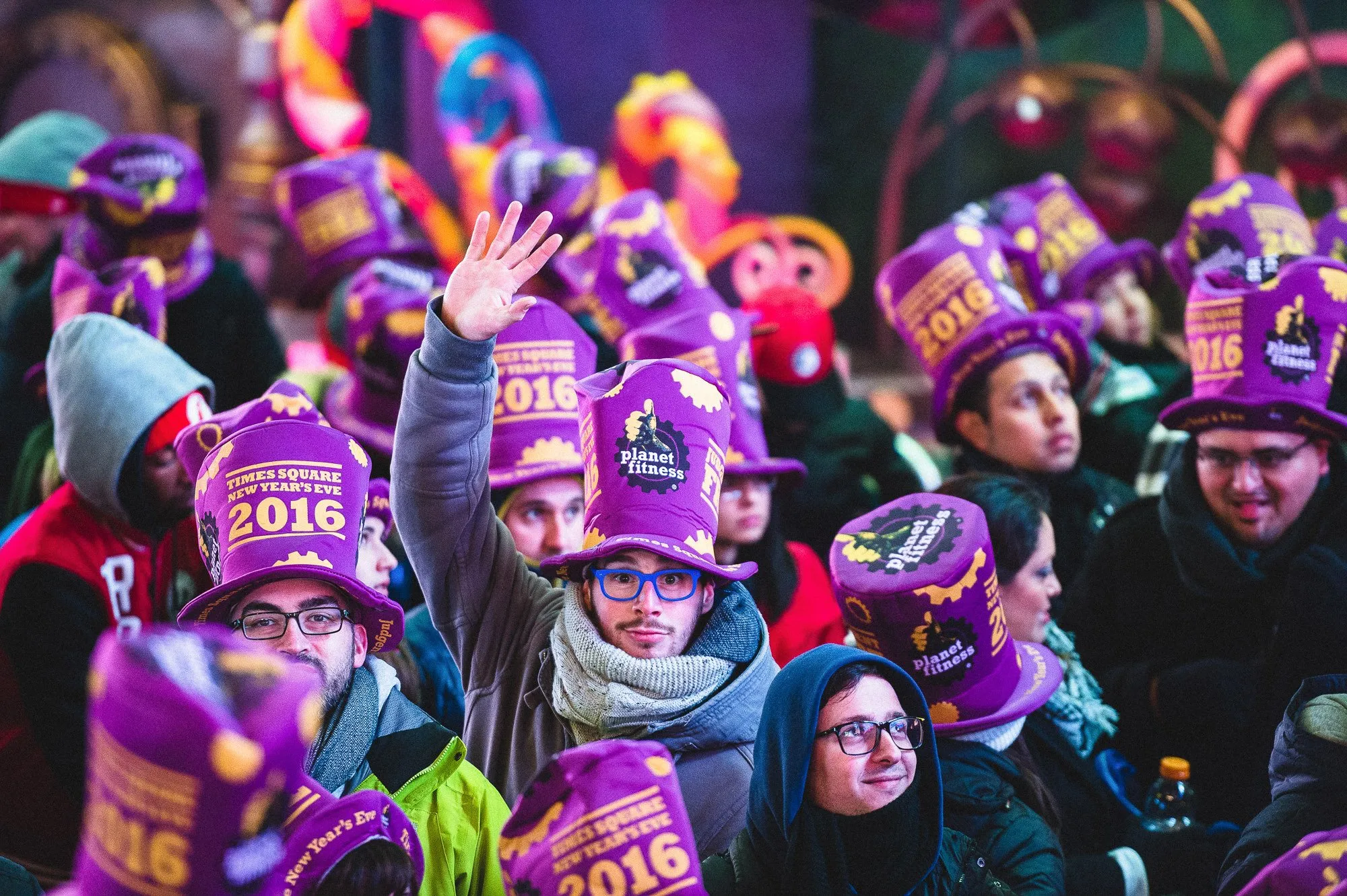 Crowd of people at Times Square New Year's Eve 2016 celebration wearing tall purple hats with 'Times Square New Year's Eve 2016' and 'Planet Fitness' logos, one person in the center raising their hand wearing blue glasses and a scarf.