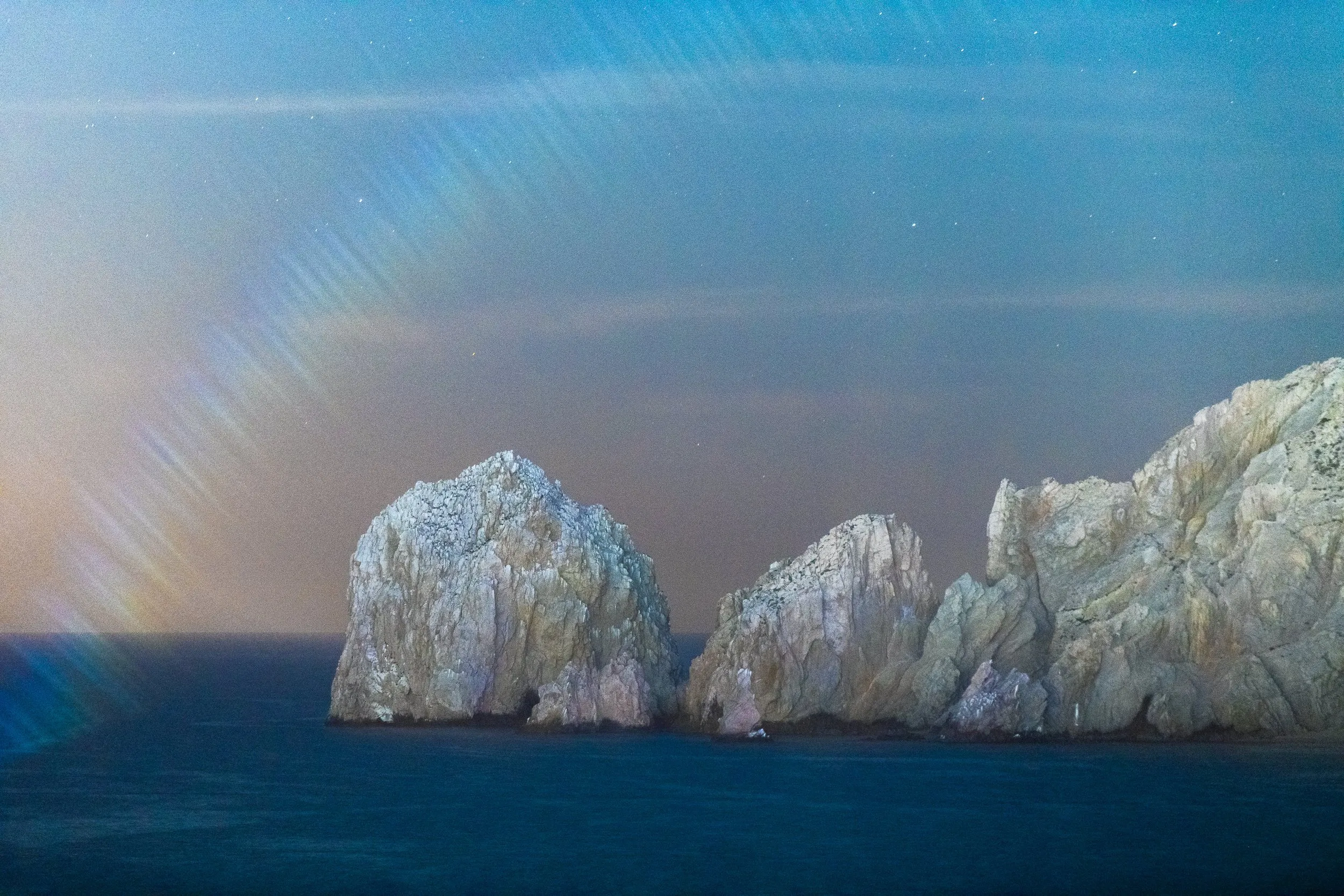 Large rugged rock formations rising from a calm dark blue sea under a twilight sky with visible stars and a faint rainbow-like light effect.