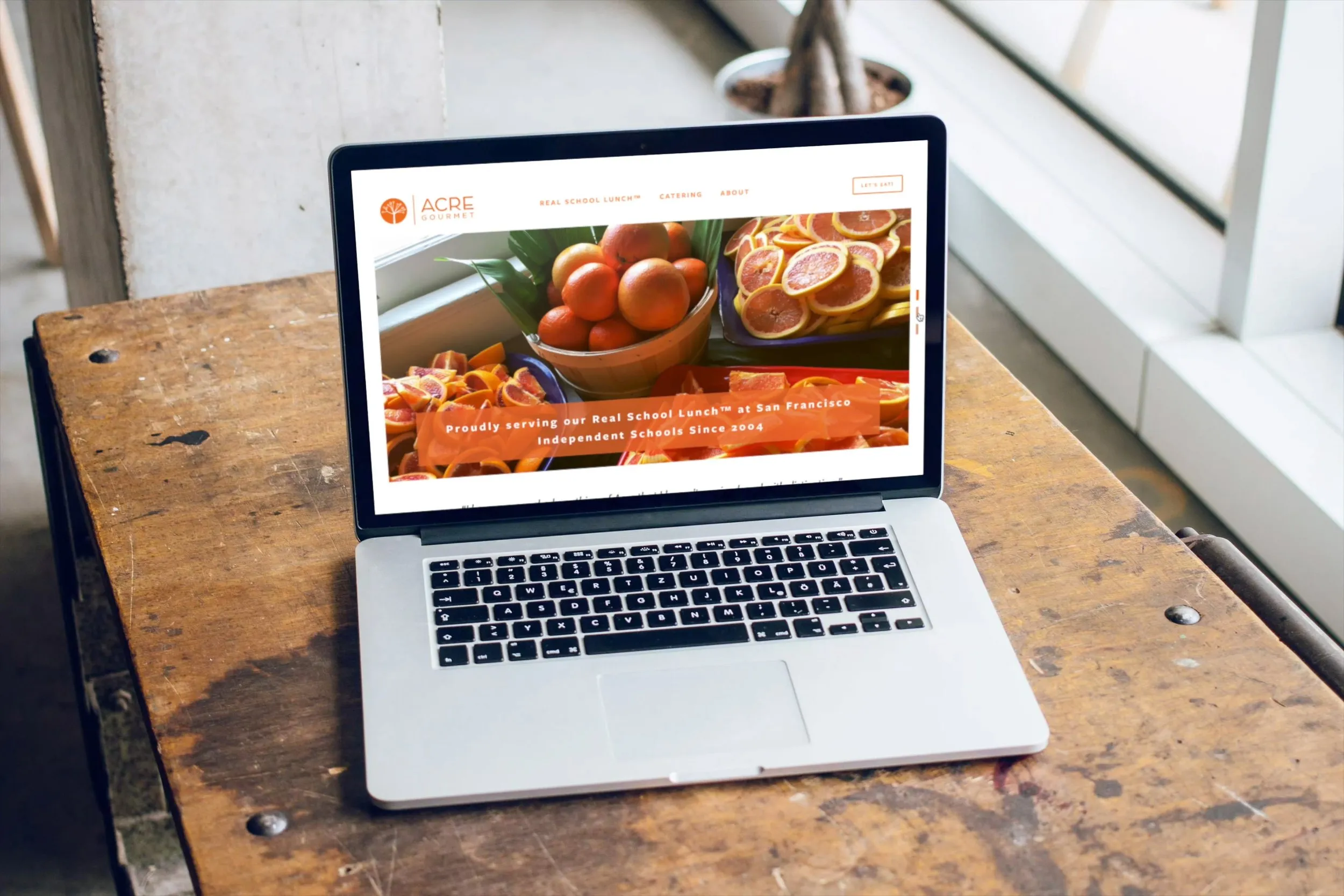 Open laptop on a wooden table displaying the Acre Gourmet website with images of fresh oranges and sliced citrus fruits, and text about serving Real School Lunch at San Francisco Independent Schools since 2004.