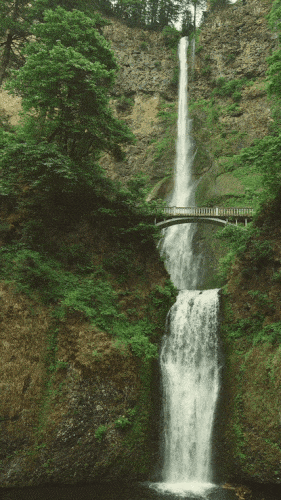 Waterfall pouring down a mossy cliff beneath a stone footbridge (animated loop)