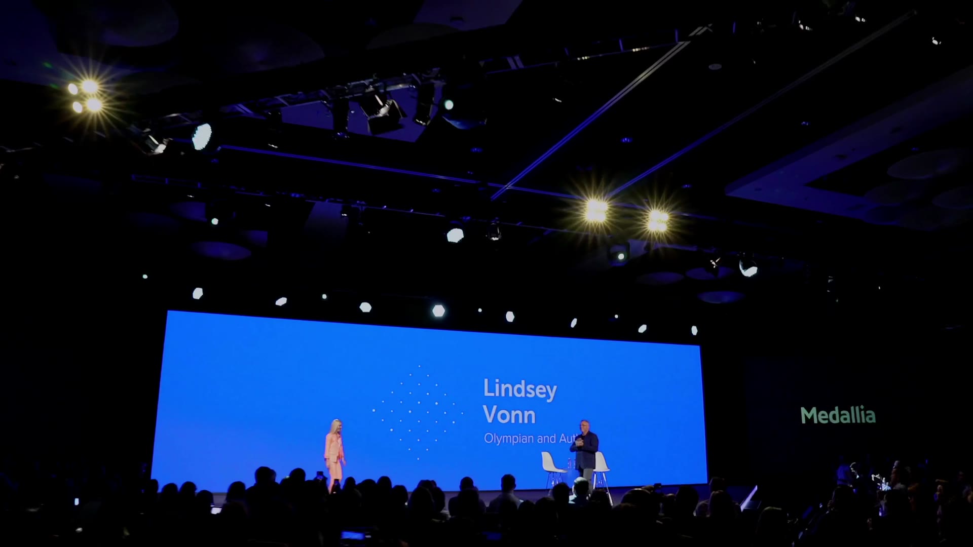 Lindsey Vonn on stage at a Medallia conference, in front of a giant blue screen and a packed audience.