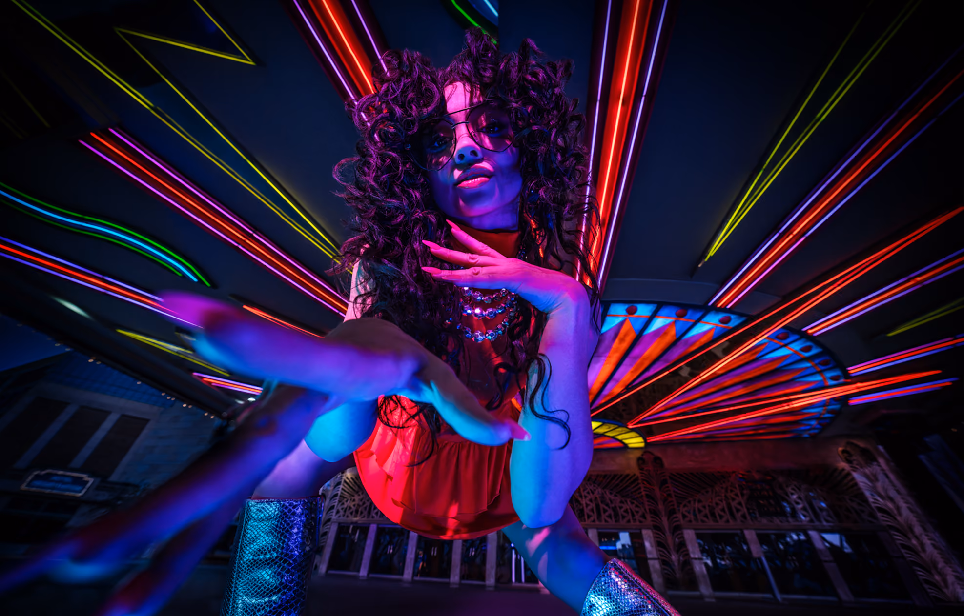 Person with curly hair wearing sunglasses, a red outfit, and shiny silver boots reaching toward the camera with neon lights radiating in the background.