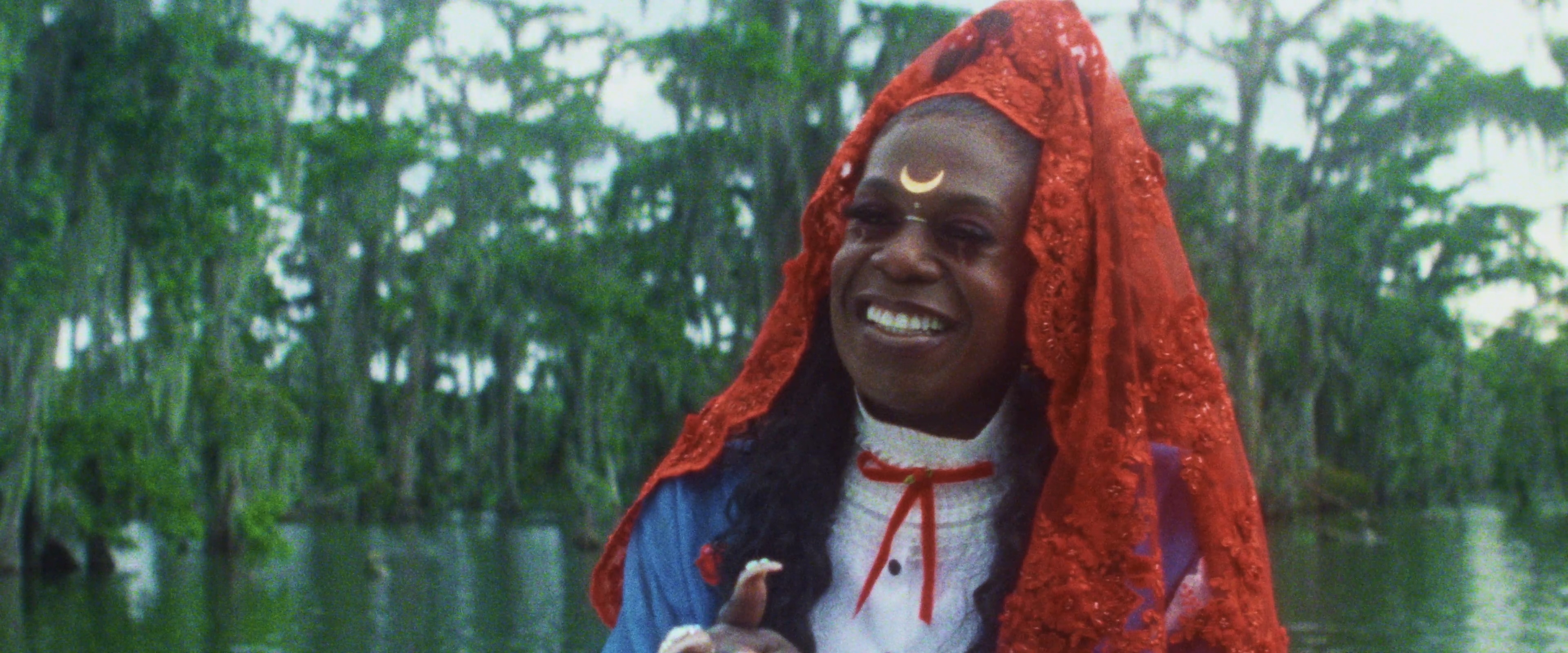 Big Freedia smiling warmly, wearing a red lace head covering and a white shirt.