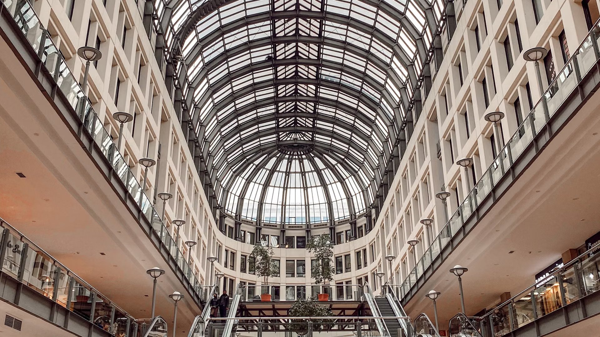 Multi-level shopping mall atrium under a glass barrel-vault roof, with escalators and retail galleries