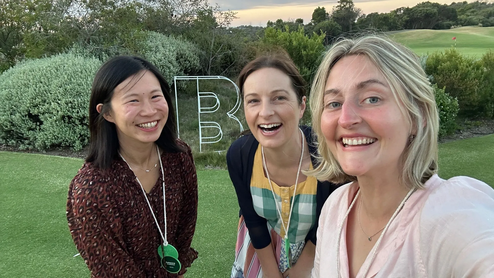 Becky, Sarah and Marnie Hawson at B Corp Assembly 2024 - Three smiling women pose for a selfie at an outdoor B Corp event, with a large “B” sign and greenery in the background.