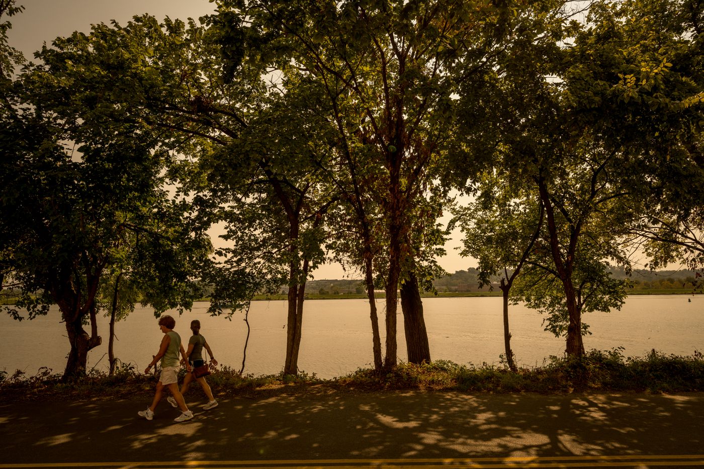 Two people walking a tree-lined riverside path at Buzzard Point Park at golden hour
