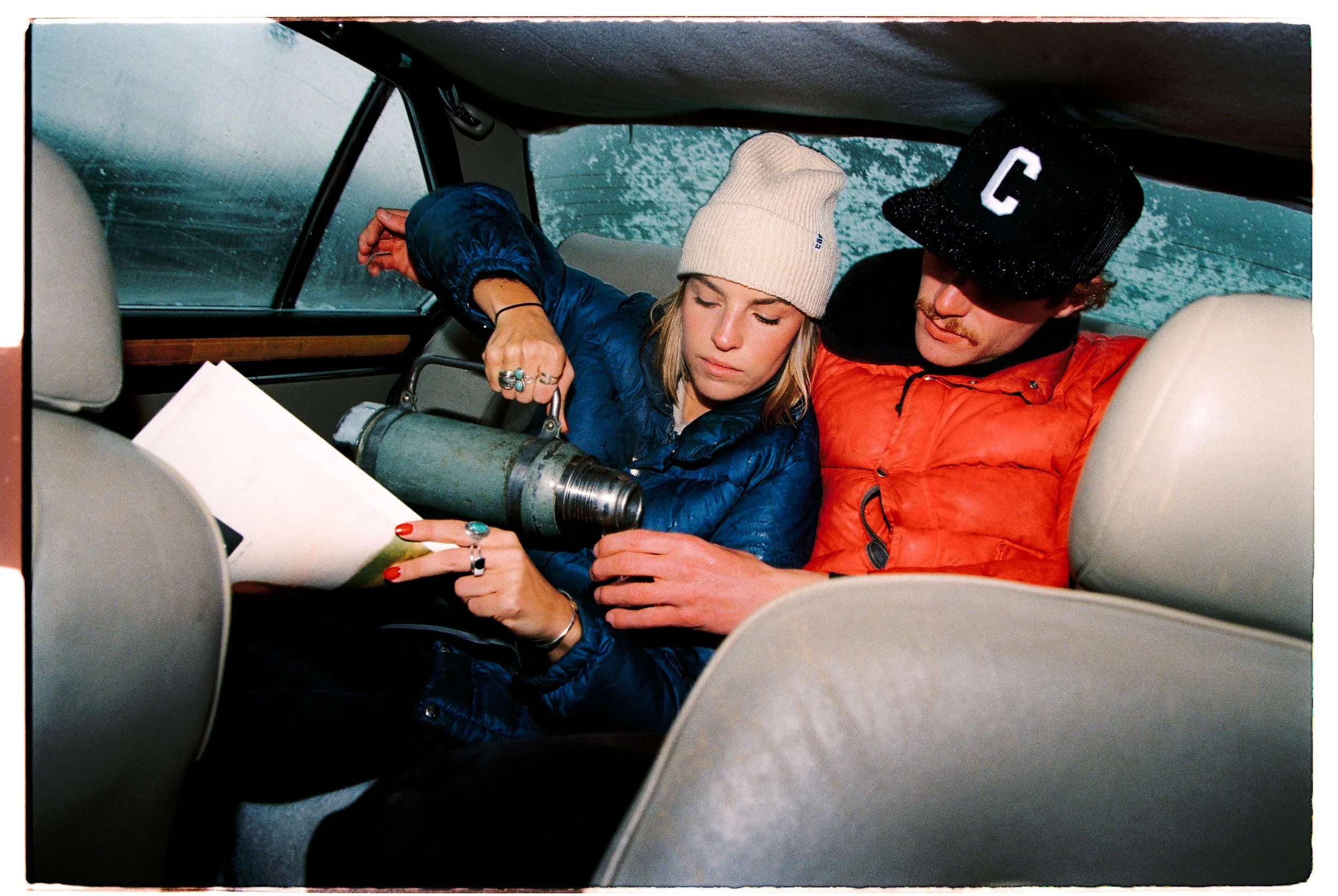 Couple in a car's back seat looking at a notebook with a vintage thermos — CAO Winter 25