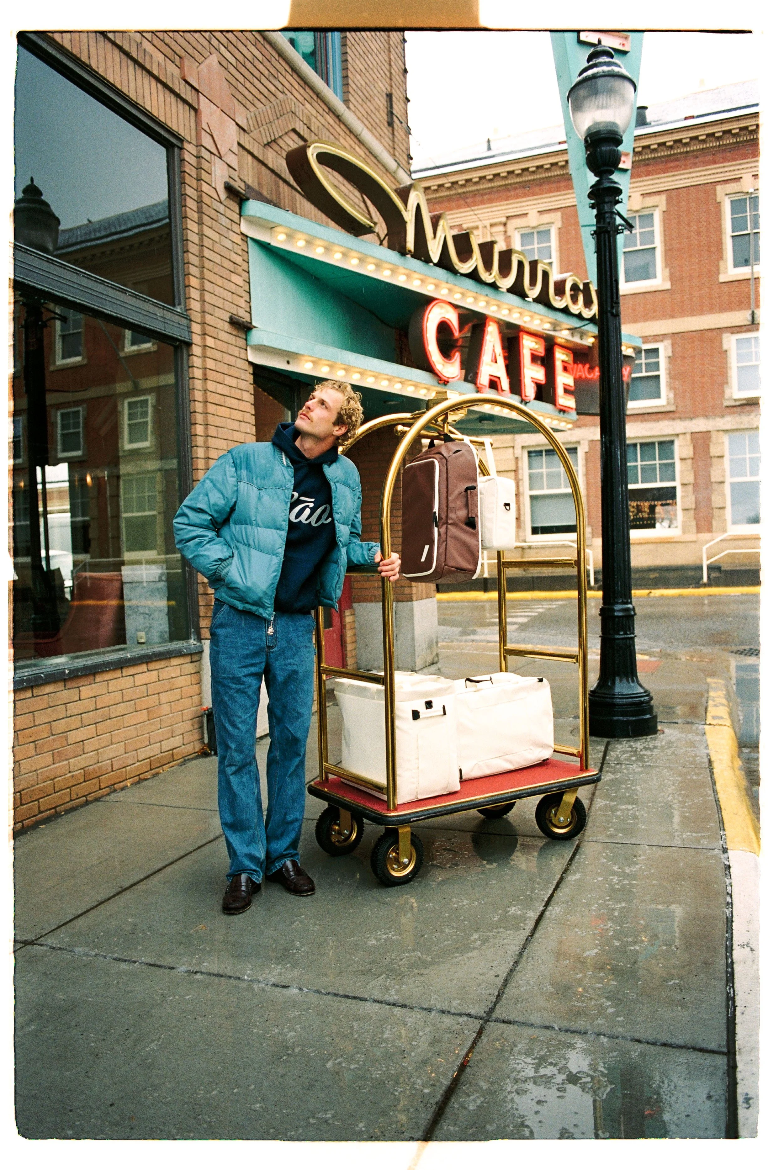 Traveler with a luggage cart outside a neon-lit café — CAO Winter 25