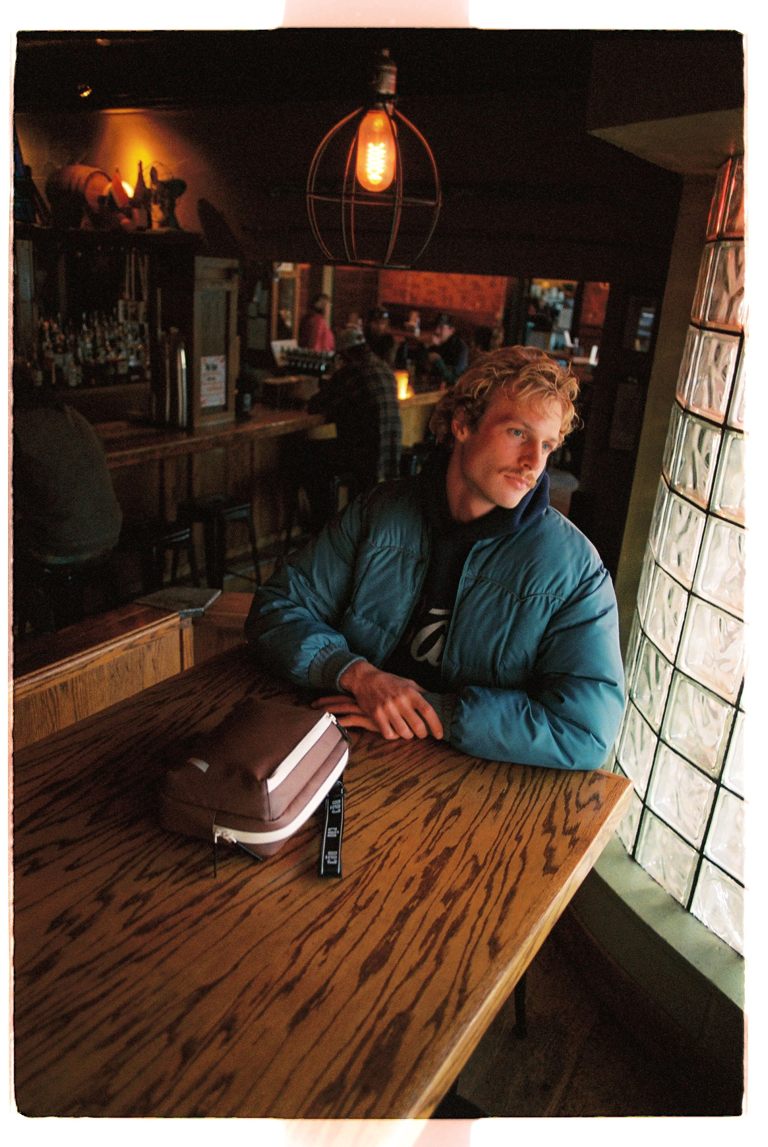 Young man in a puffer jacket alone at a café table by the window — CAO Winter 25