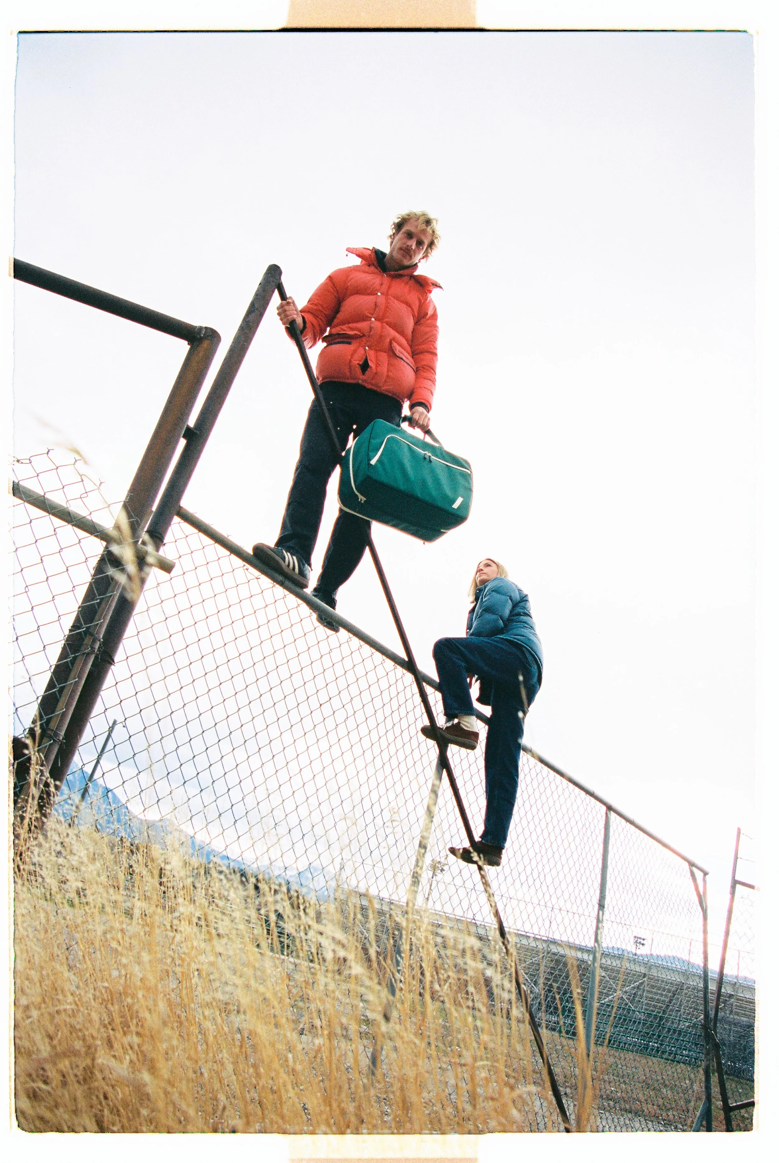 Two travelers climbing a chain-link fence with a duffel bag — CAO Winter 25