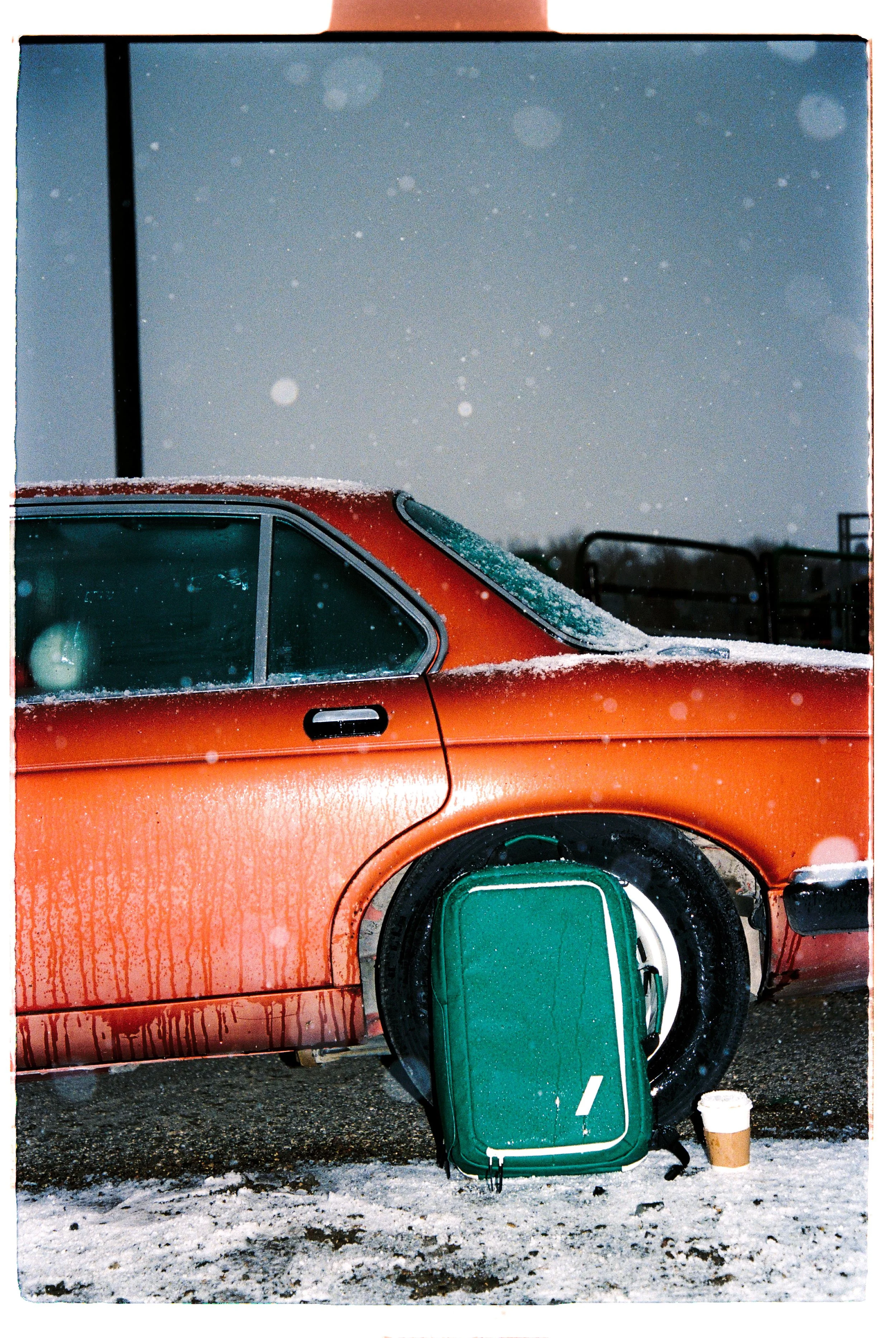 Snow-covered orange car with a green suitcase against the tire — CAO Winter 25