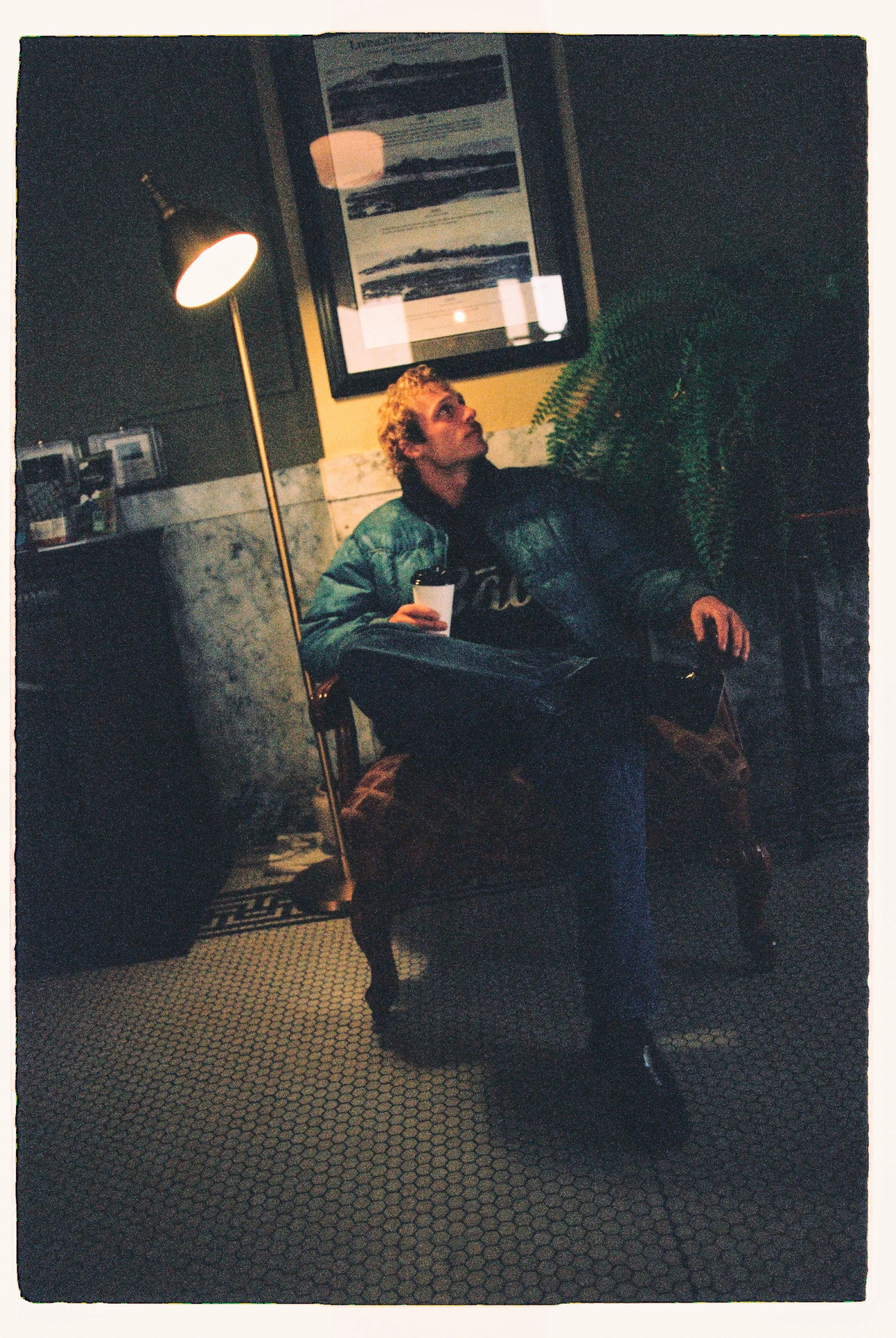 Traveler in a green puffer jacket holding a cup in a dim café — CAO Winter 25