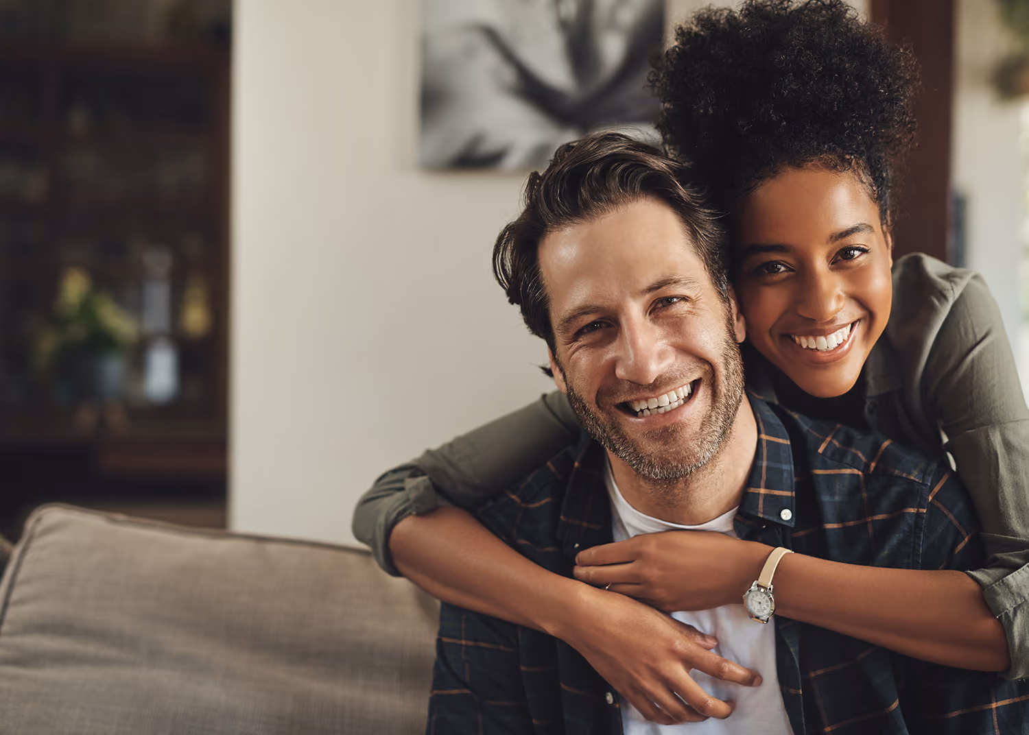Smiling couple sitting on a couch with the woman hugging the man from behind in a cozy indoor setting.