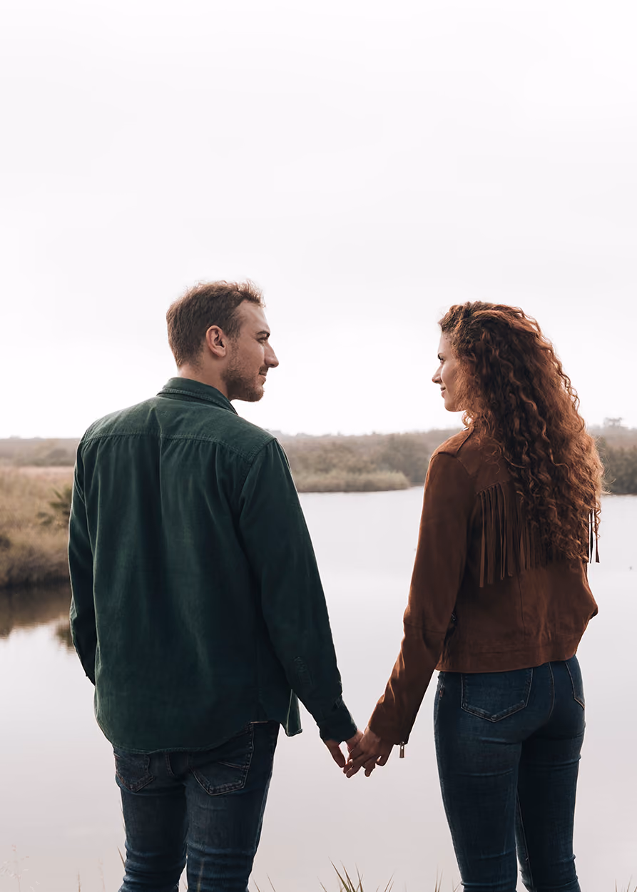 Man and woman holding hands and looking at each other while standing near a calm river with shrubs and trees in the background under an overcast sky.