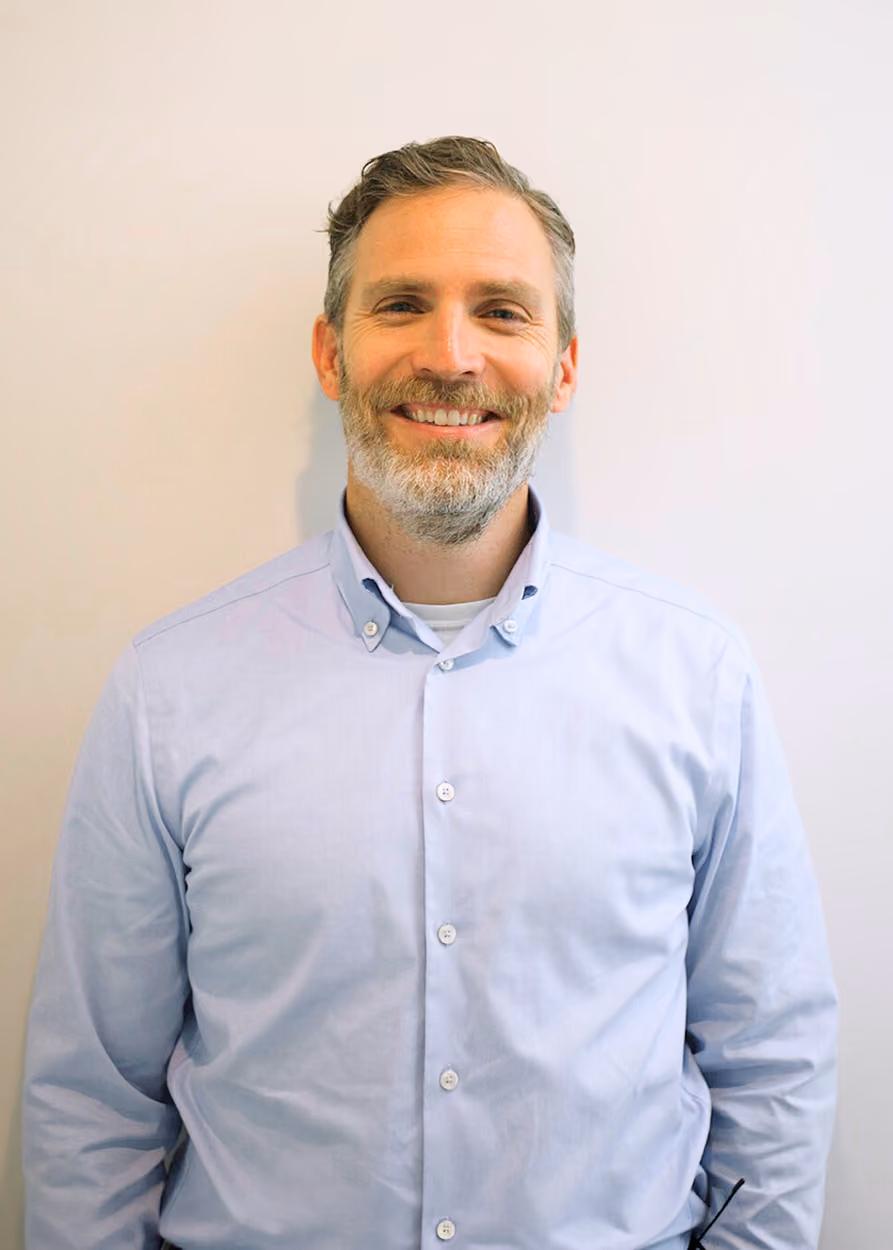 Smiling middle-aged man with light skin, short grayish hair, and a beard wearing a light blue button-up shirt standing against a plain light background.