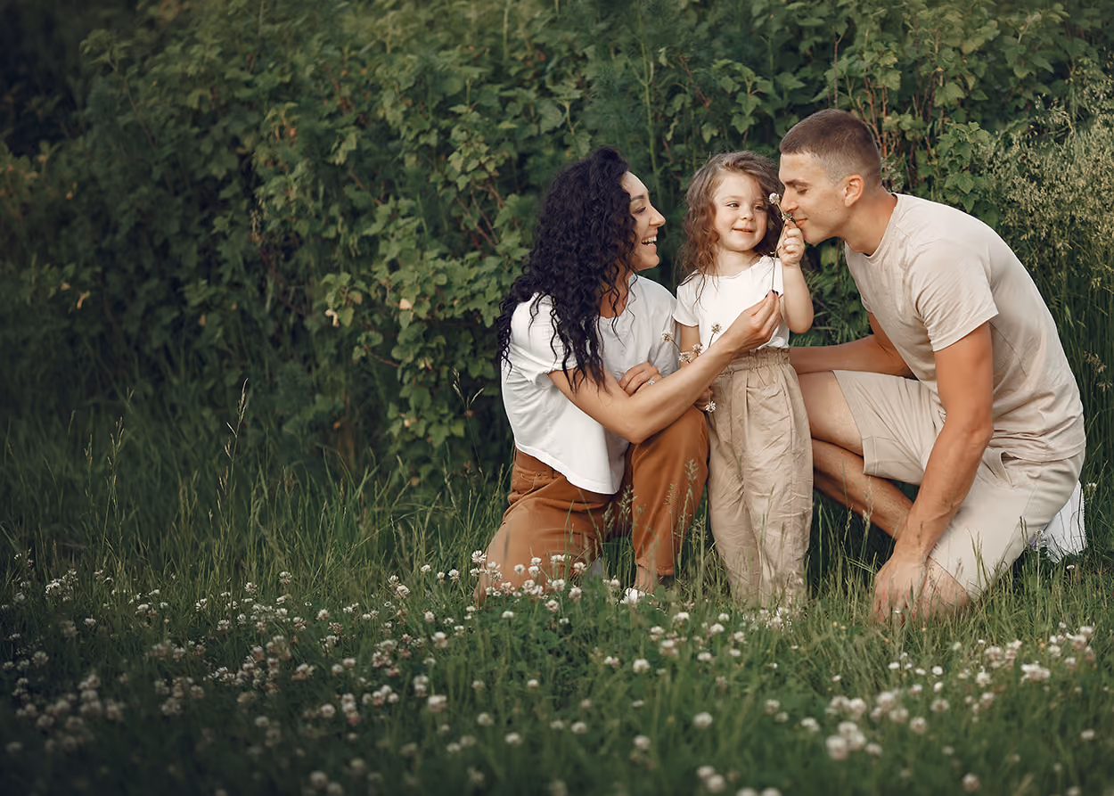A smiling family outdoors with a woman and a man kneeling on grass beside a young girl holding a small flower, surrounded by greenery and wildflowers.