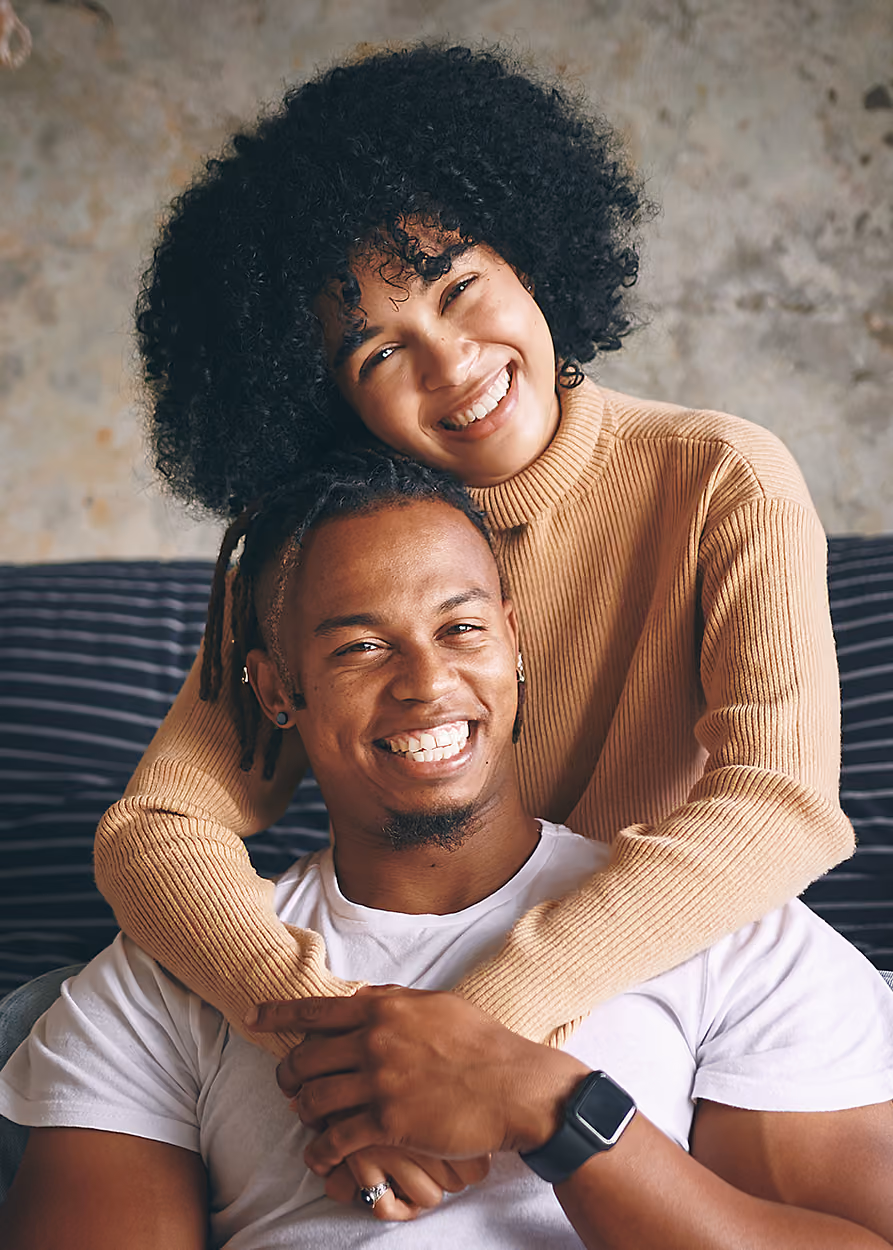 Smiling couple indoors; woman with curly hair wearing beige sweater hugs man with short dreadlocks wearing white t-shirt and smartwatch while sitting on a sofa.