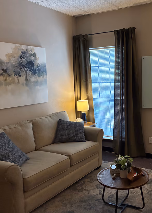 Cozy room with a beige sofa adorned with two dark cushions, a small wooden round table with a potted plant and items on it, a side table with a lamp, dark curtains partially covering a window with blinds, and a painting of trees on the wall.