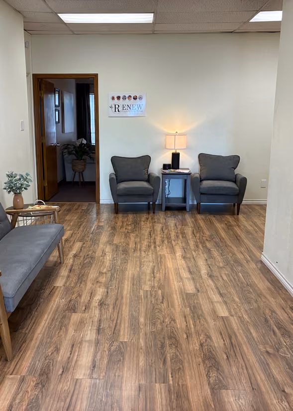 Waiting area with wooden floor, two gray armchairs separated by a small table holding a lamp, and a gray bench with a small plant on a side table. There is an open wooden door leading to another room with a plant inside, and a sign on the wall behind the chairs.