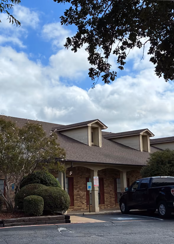 Brick building with a brown roof and two dormer windows under a partly cloudy blue sky, surrounded by trees and bushes, with a black pickup truck parked in front on a paved lot.