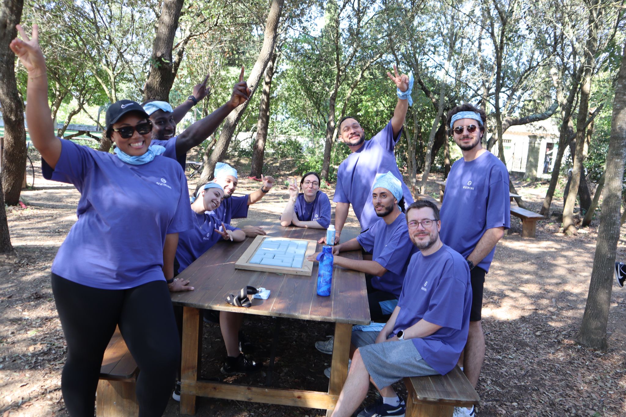 Un groupe de neuf personnes portant des t-shirts violets et des bandanas bleus posent joyeusement autour d'une table en bois dans une forêt ensoleillée, certains faisant des signes de paix avec les mains.