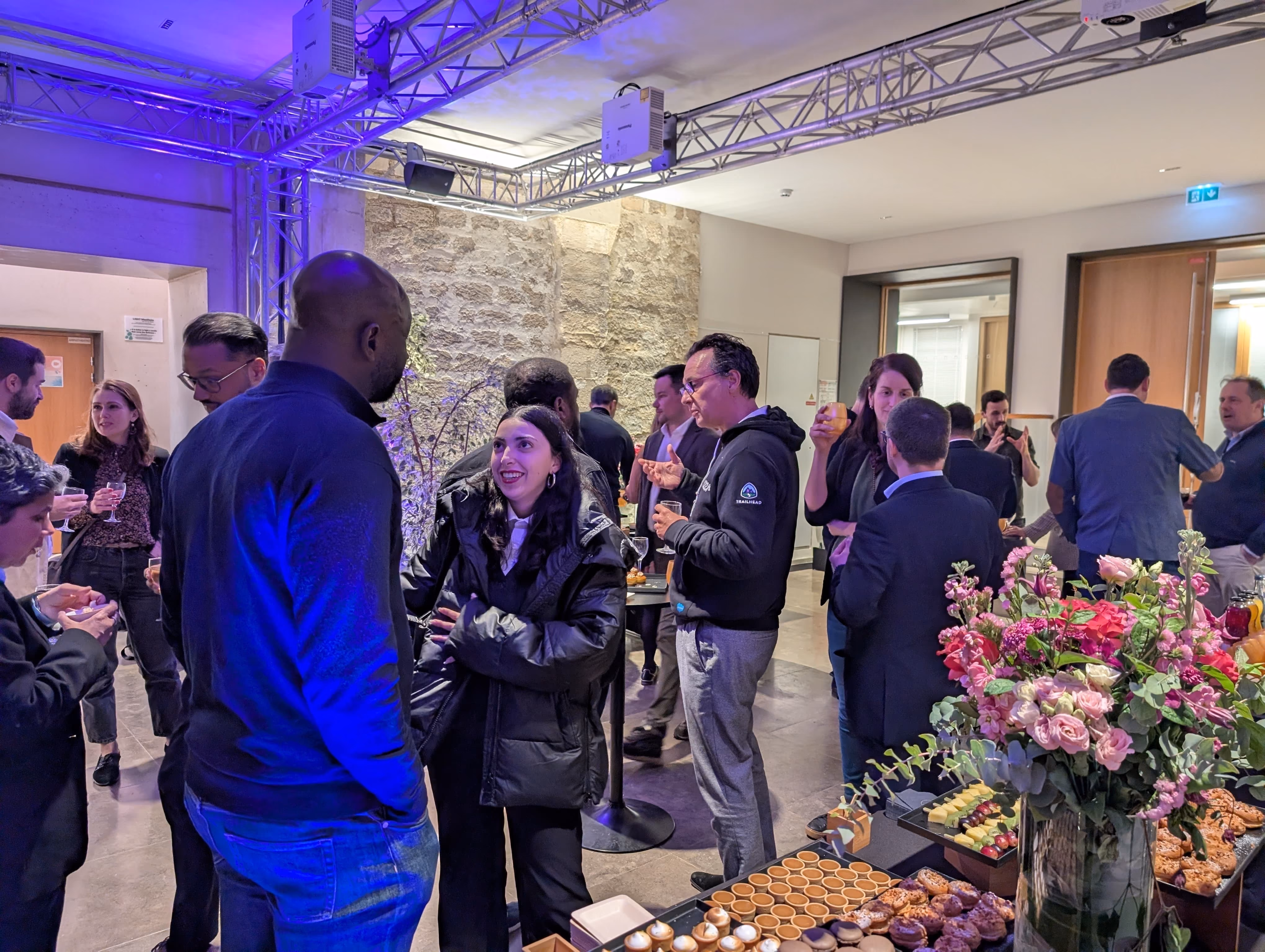 Groupe de personnes discutant lors d'un événement social en intérieur avec éclairage violet, autour de tables hautes avec des boissons et un buffet de pâtisseries, avec un grand vase de fleurs colorées au premier plan.