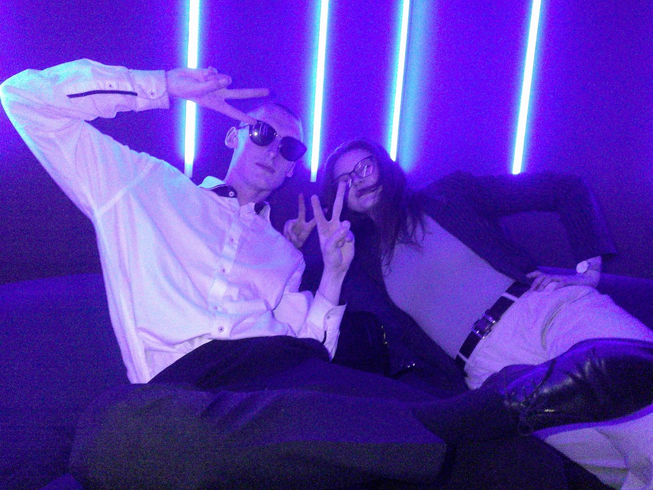 Mężczyzna w białej koszuli i kobieta w okularach pokazują znak pokoju w pokoju Karaoke Spot z fioletowym neonowym światłem