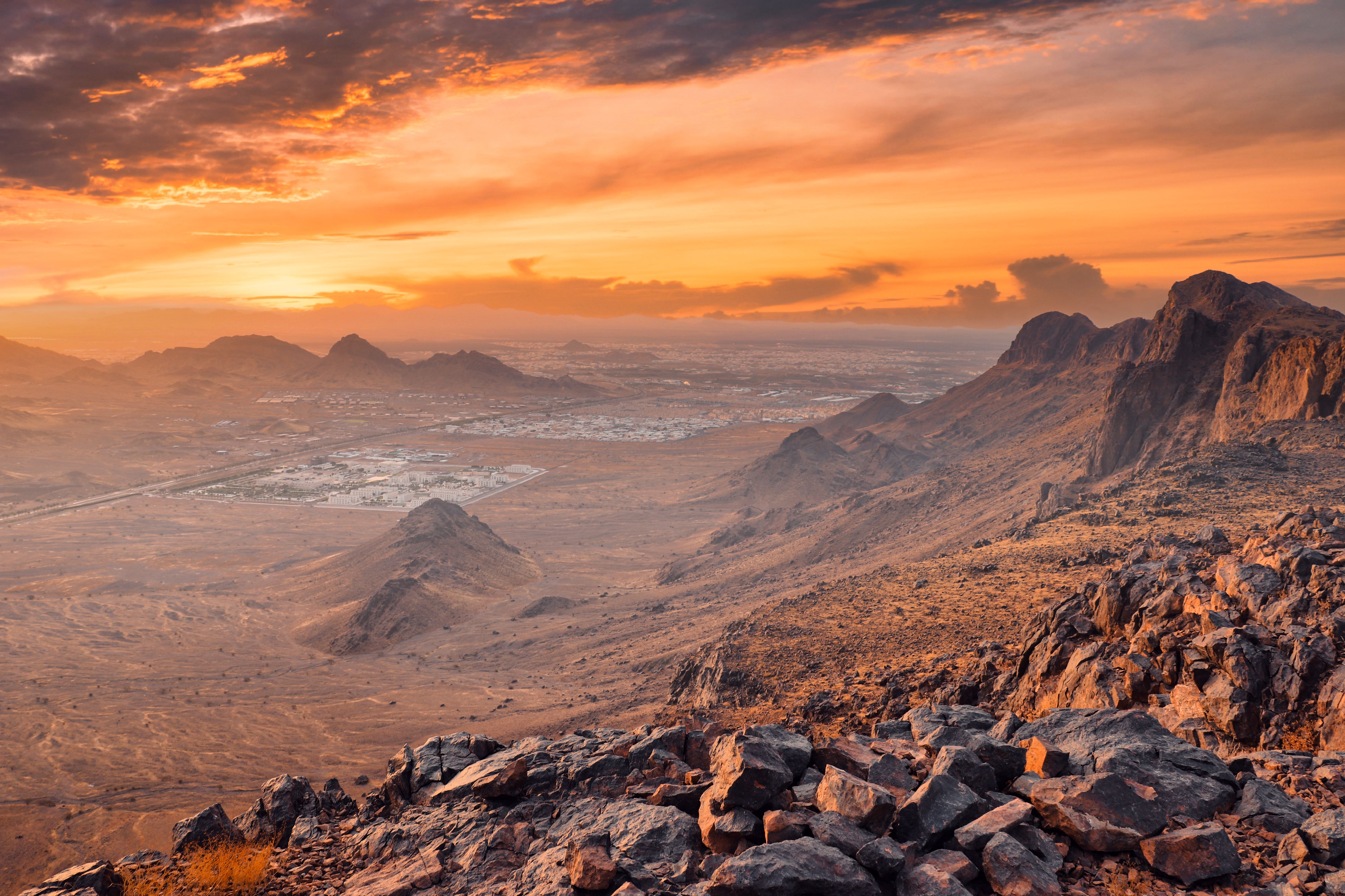 Rocky mountain landscape at sunset with an orange sky, rugged peaks, and a dry valley featuring sparse vegetation and a distant settlement.