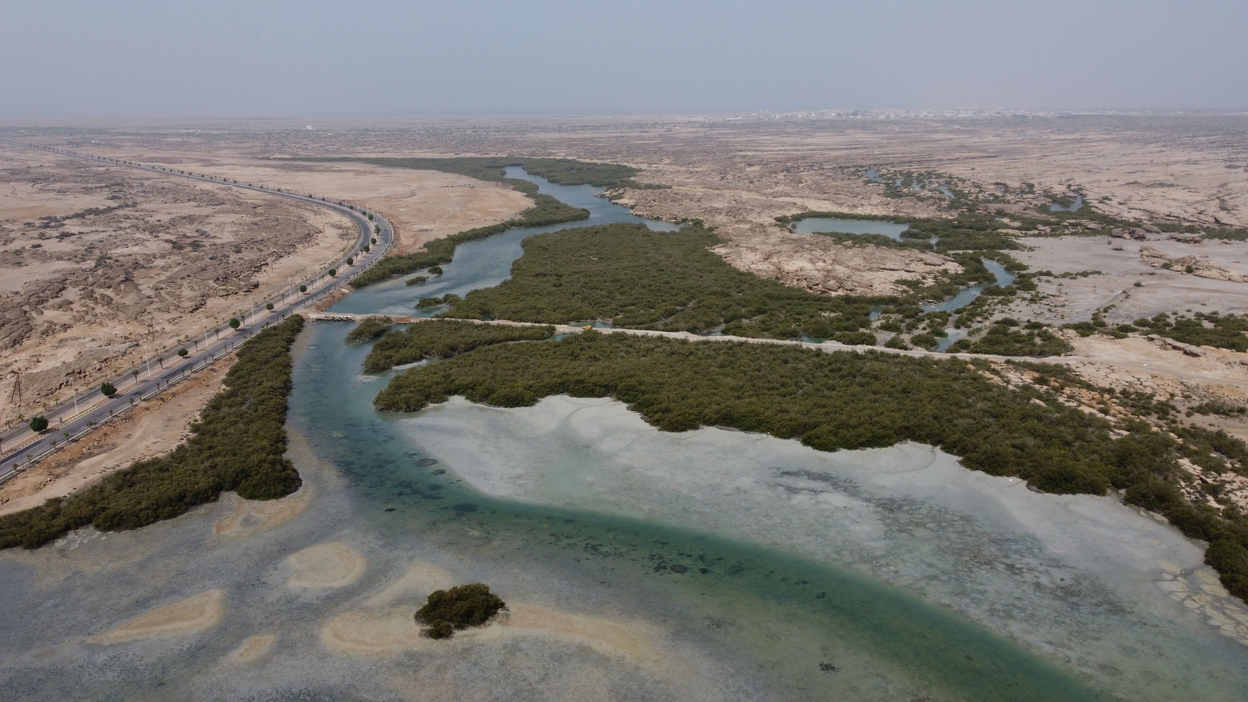 Aerial view of a river winding through a dry, rocky landscape with dense green mangroves along its banks and a paved road parallel to the river on the left side.
