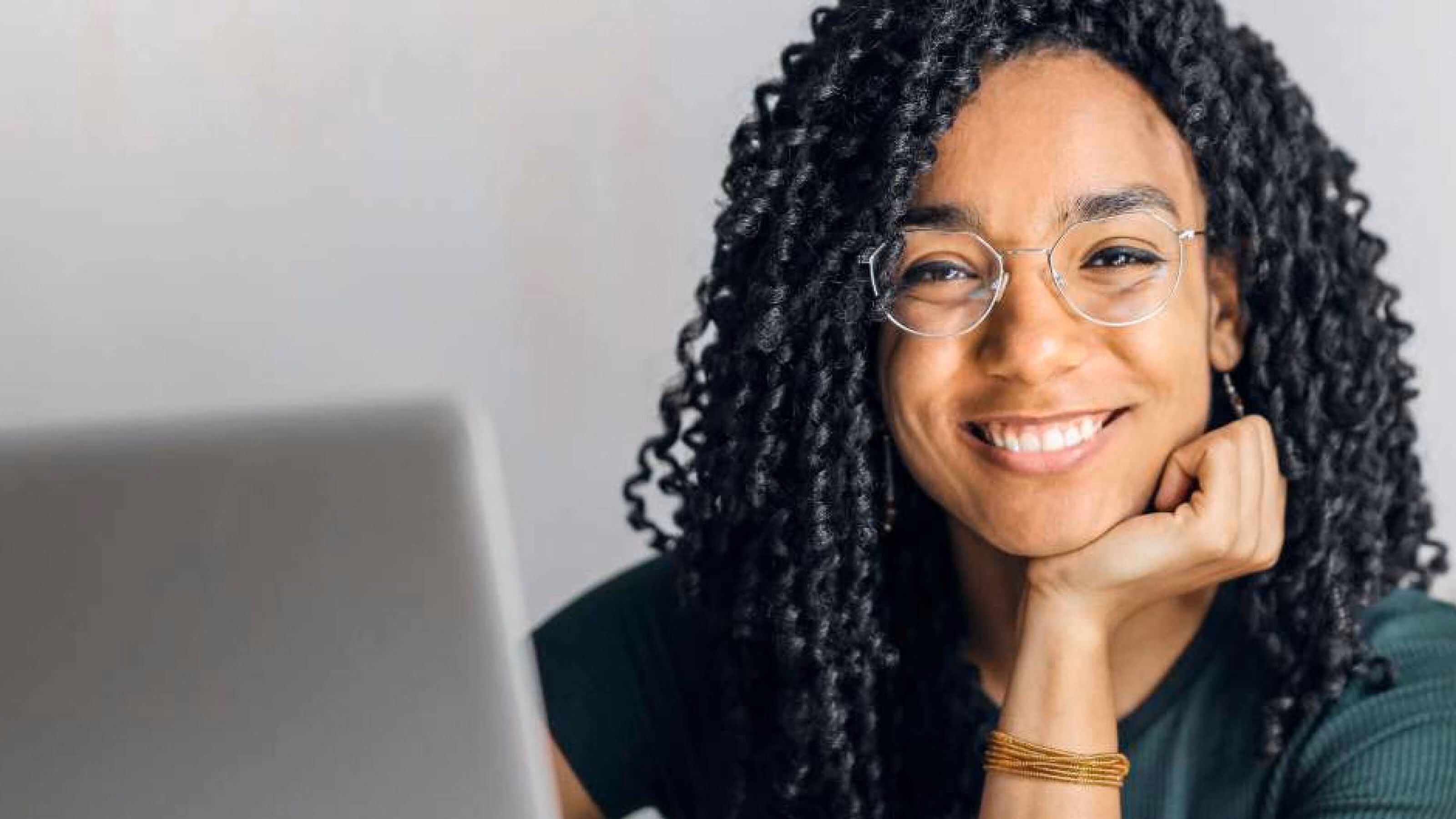 Smiling woman with curly black hair and glasses resting her chin on her hand, sitting near a laptop.