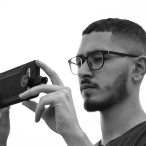 Black and white photo of a man with glasses and a beard holding a camera up to his eye
