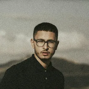 Portrait of a young man with short dark hair, a beard and glasses, wearing a black collared shirt