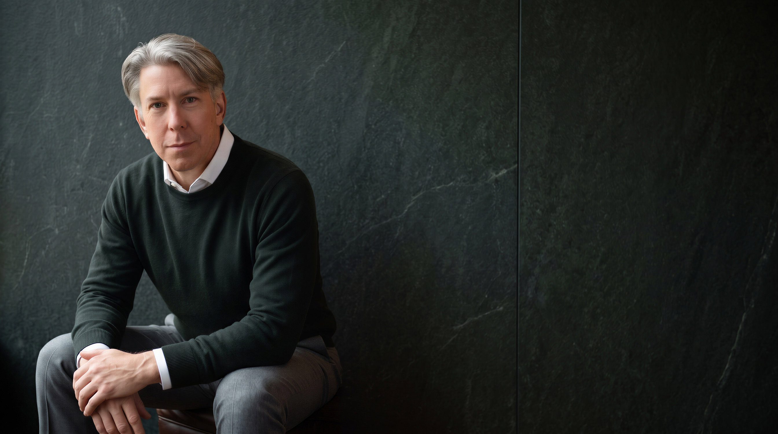 Dan Reed, seated and leaning forward against a dark slate wall.