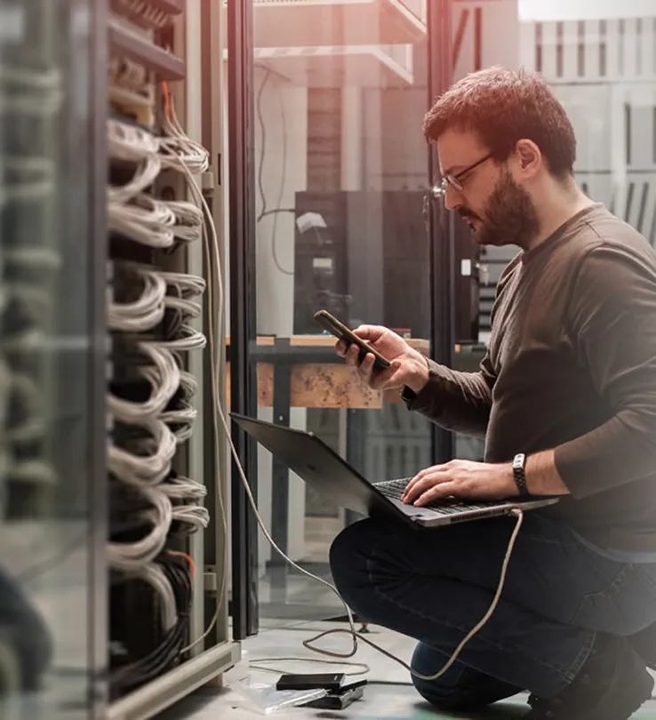 A technician checking a network rack on a laptop and phone