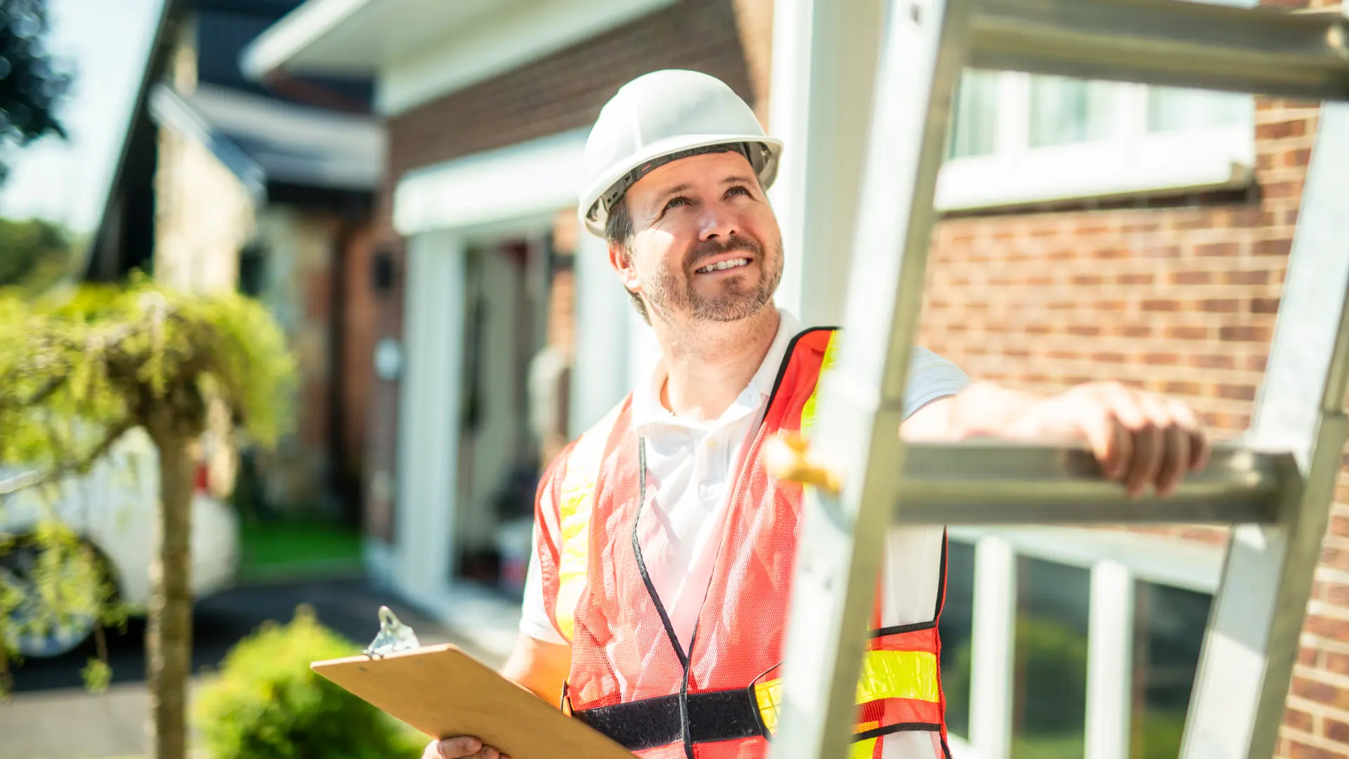 A public adjuster in a hard hat and safety vest looking up at a home's roofline beside a ladder, clipboard in hand