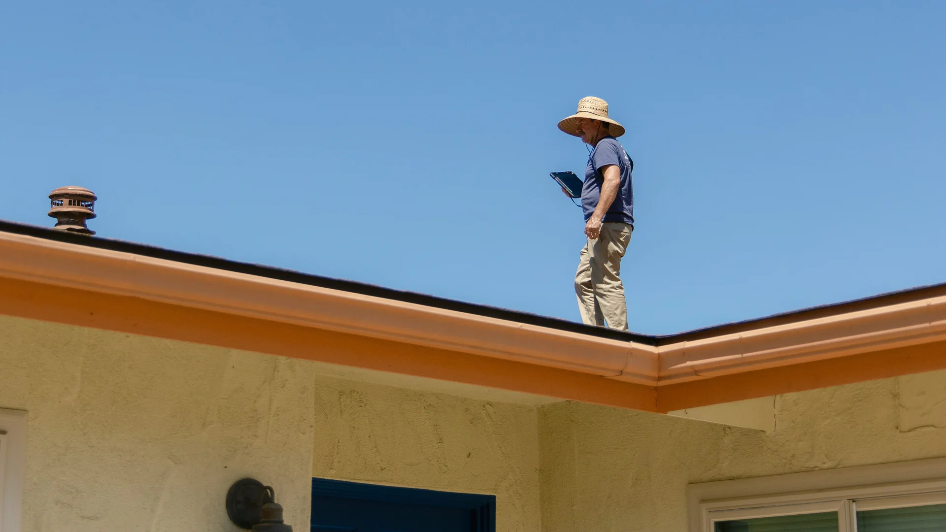 A public adjuster on a rooftop in a sun hat, checking his inspection notes on a tablet