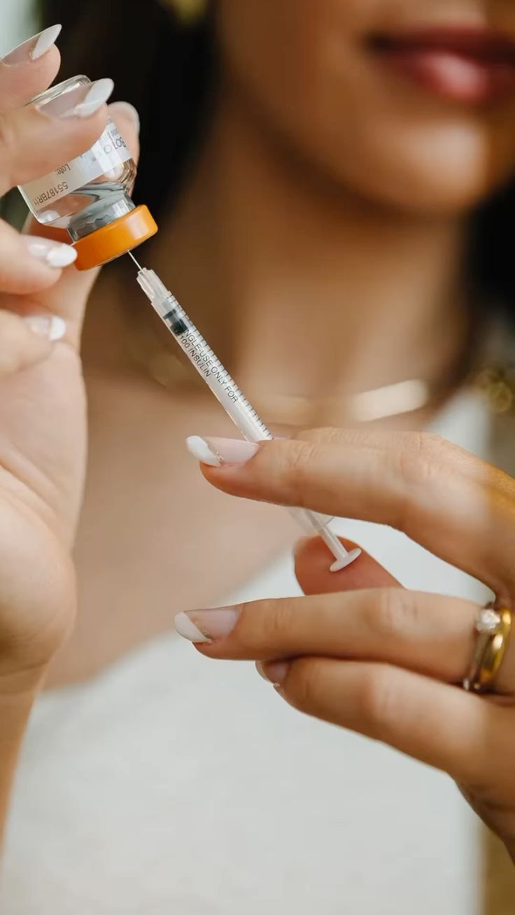 Close-up of a person drawing liquid from a vial with an orange cap into an insulin syringe, showing hands with manicured nails and a ring.