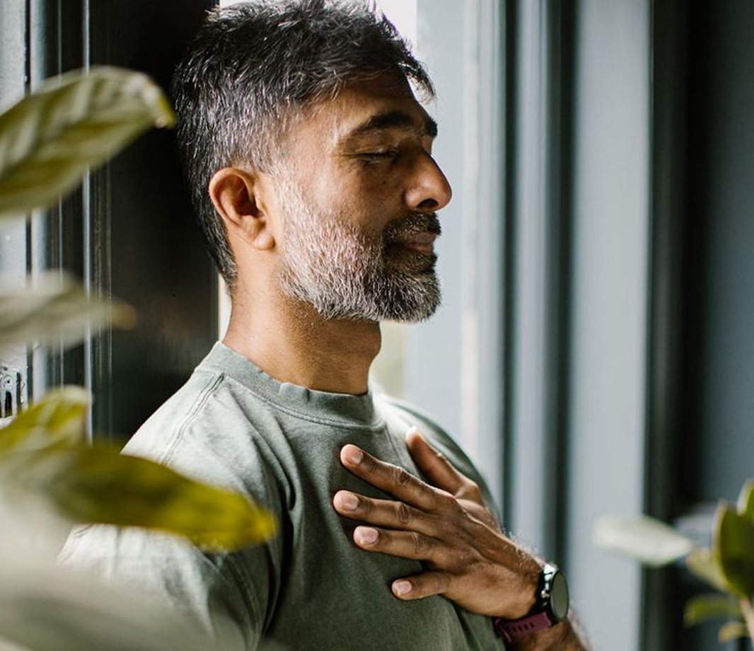 A man with a grey beard standing by a window, hand on his chest, eyes closed