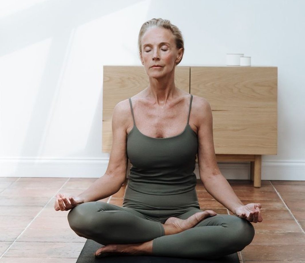 A woman meditating cross-legged on a mat at home, eyes closed