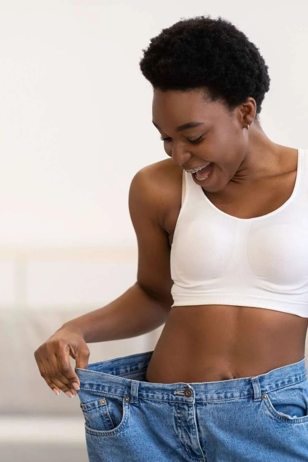 Smiling woman wearing a white sports bra holds out the waistband of oversized blue jeans to show weight loss.