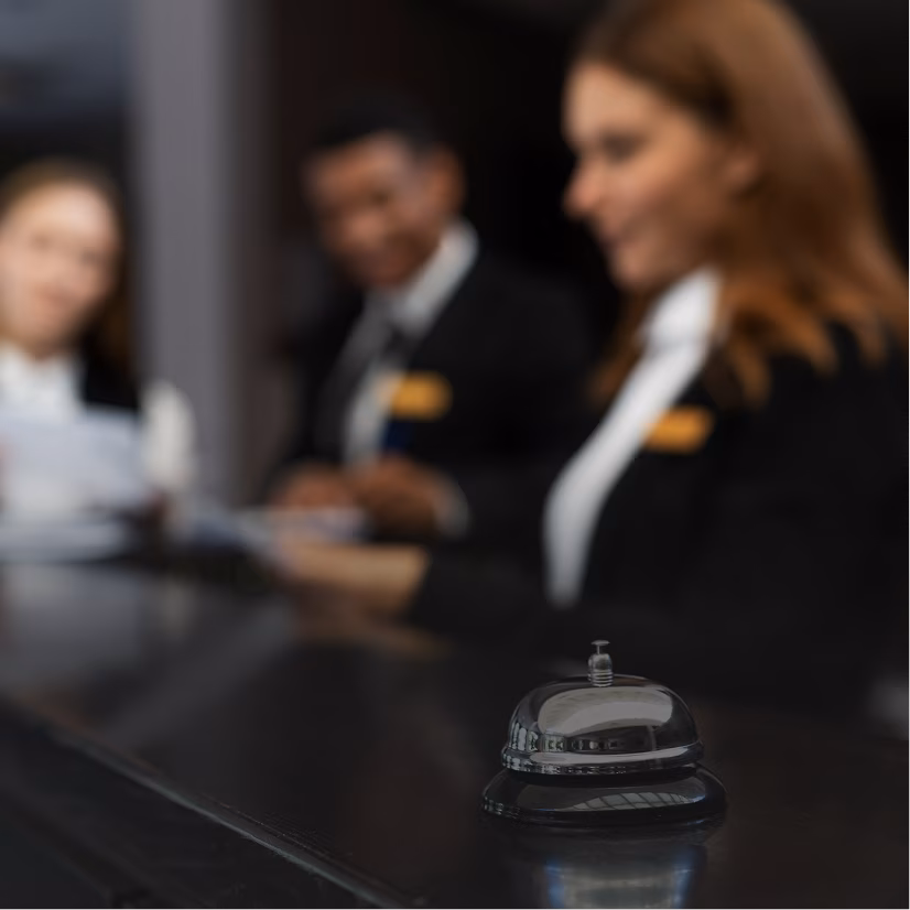 Shiny service bell on a dark countertop with three blurred hotel staff in uniform behind it.