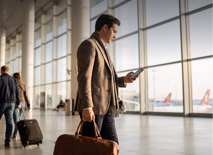 Man in a brown blazer and scarf standing in an airport terminal holding a brown leather bag and looking at his smartphone, with large windows and airplane tails visible outside.