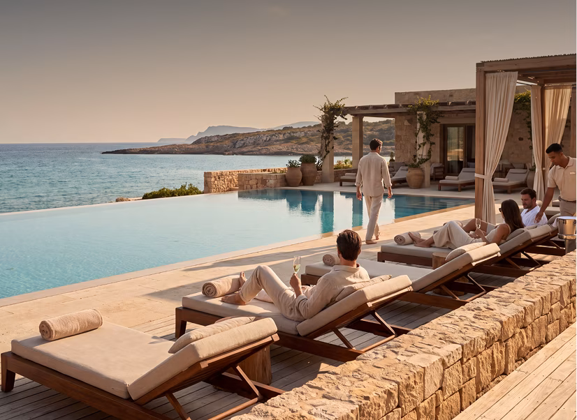 People relaxing on cushioned lounge chairs by an infinity pool overlooking a calm sea with rocky coastline, next to a stone patio and modern building with outdoor seating and curtains.