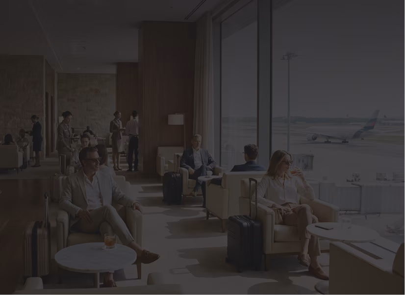 Passengers seated in a modern airport lounge with large windows overlooking the airfield where a plane is visible, some people are sitting with luggage while others stand and converse.